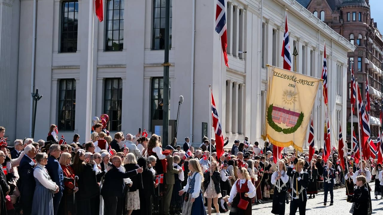 Folk i bunad går i 17. mai-tog med norske flagg og en gul fane foran Universitetet i Oslo, med publikum langs gata